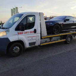 Biała laweta Peugeot z numerem telefonu i logo firmy holowniczej przewozi czarne BMW na tle parkingu i szyld&oacute;w McDonald's o zmierzchu.