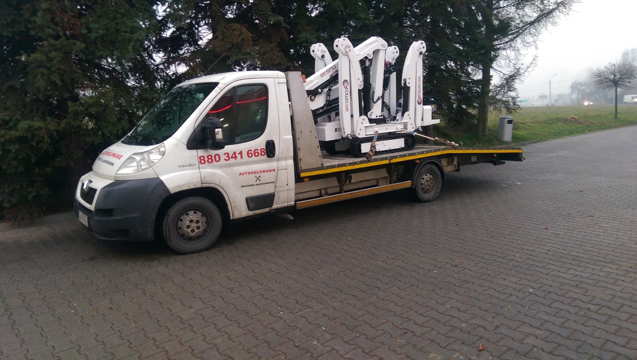 Biała laweta Peugeot z platformą przewożąca małą białą koparkę gąsienicową z ramionami, zaparkowana na brukowanym parkingu obok drzew.