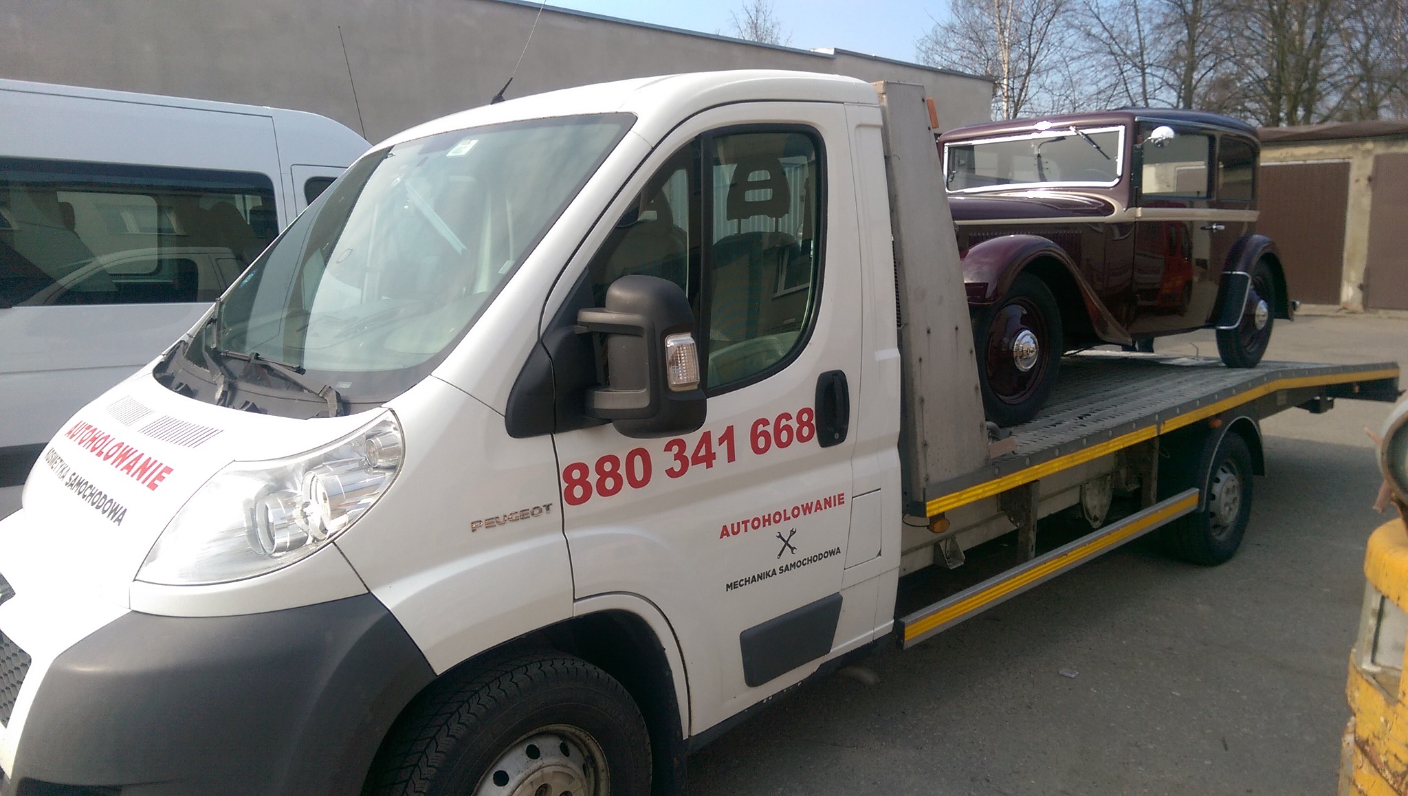 Biała laweta Peugeot z czerwonym numerem telefonu i logo firmy, przewożąca zabytkowy, ciemnoczerwony samochód osobowy z chromowanymi elementami.