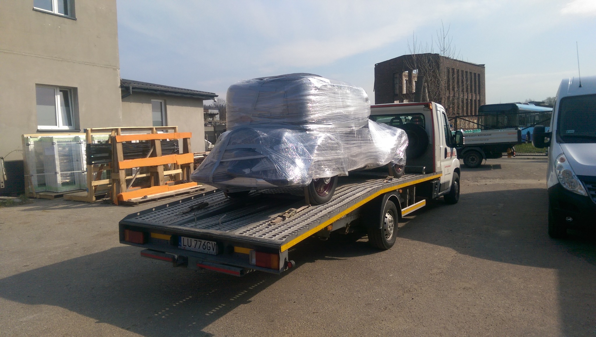Samochód zabytkowy owinięty folią transportową, przewożony na platformie lawety, z widocznymi paletami z oknami w tle.