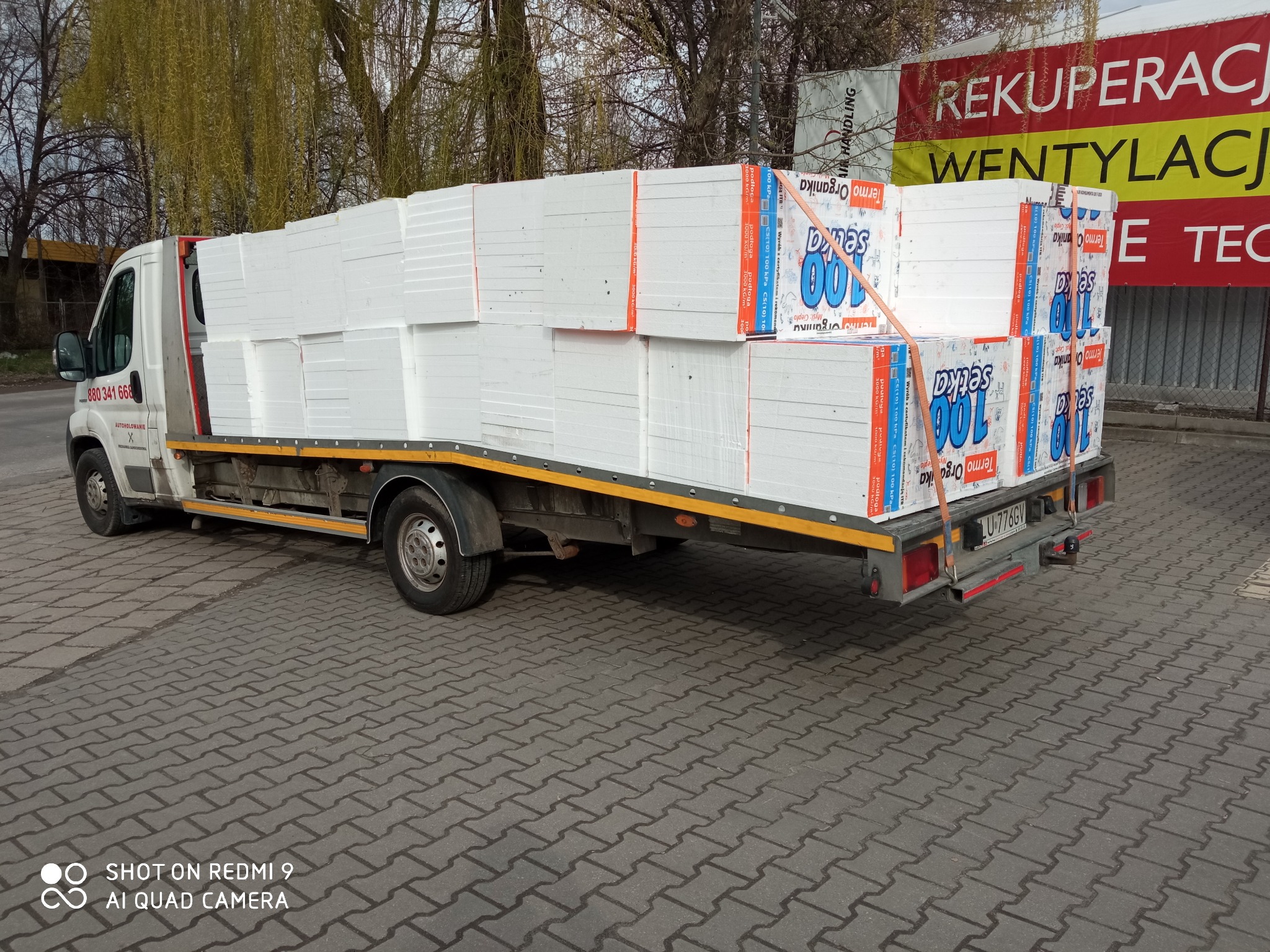 Biały bus z platformą załadowany paletami styropianu Termo Organika w Lublinie, zabezpieczony pomarańczowymi pasami transportowymi, na tle budynku z reklamą rekuperacji i wentylacji.