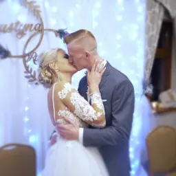 Nowożeńcy całują się na tle dekoracji ślubnej z imionami, w tle mężczyzna w garniturze.