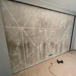 Freedom - Ściana w pokoju po nałożeniu szarej, dekoracyjnej faktury z geometrycznym wzorem z białych linii. Widoczne gniazdko elektryczne, narzędzia i resztki materiałów na podłodze.