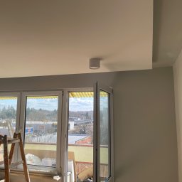 Freedom - Wnętrze mieszkania w trakcie malowania, widoczna drabina przy oknie balkonowym, sufit pomalowany na biało, ściany w odcieniach szarości i beżu, zamontowane lampy sufitowe.