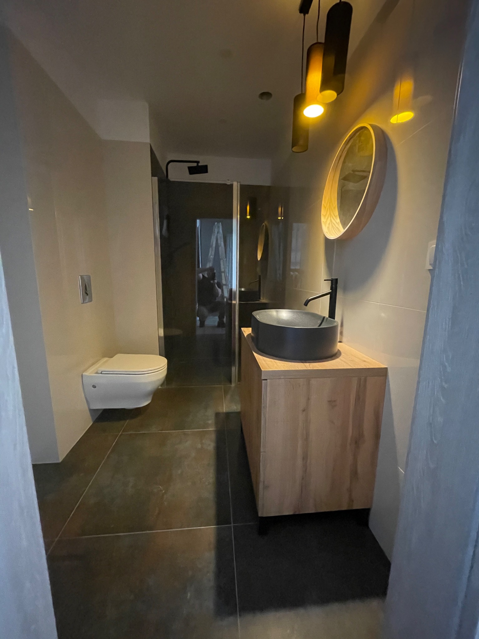 Nowoczesna łazienka z podwieszaną toaletą, umywalką nablatową w kolorze grafitowym i drewnianą szafką, oświetlona wiszącymi lampami w kształcie tub