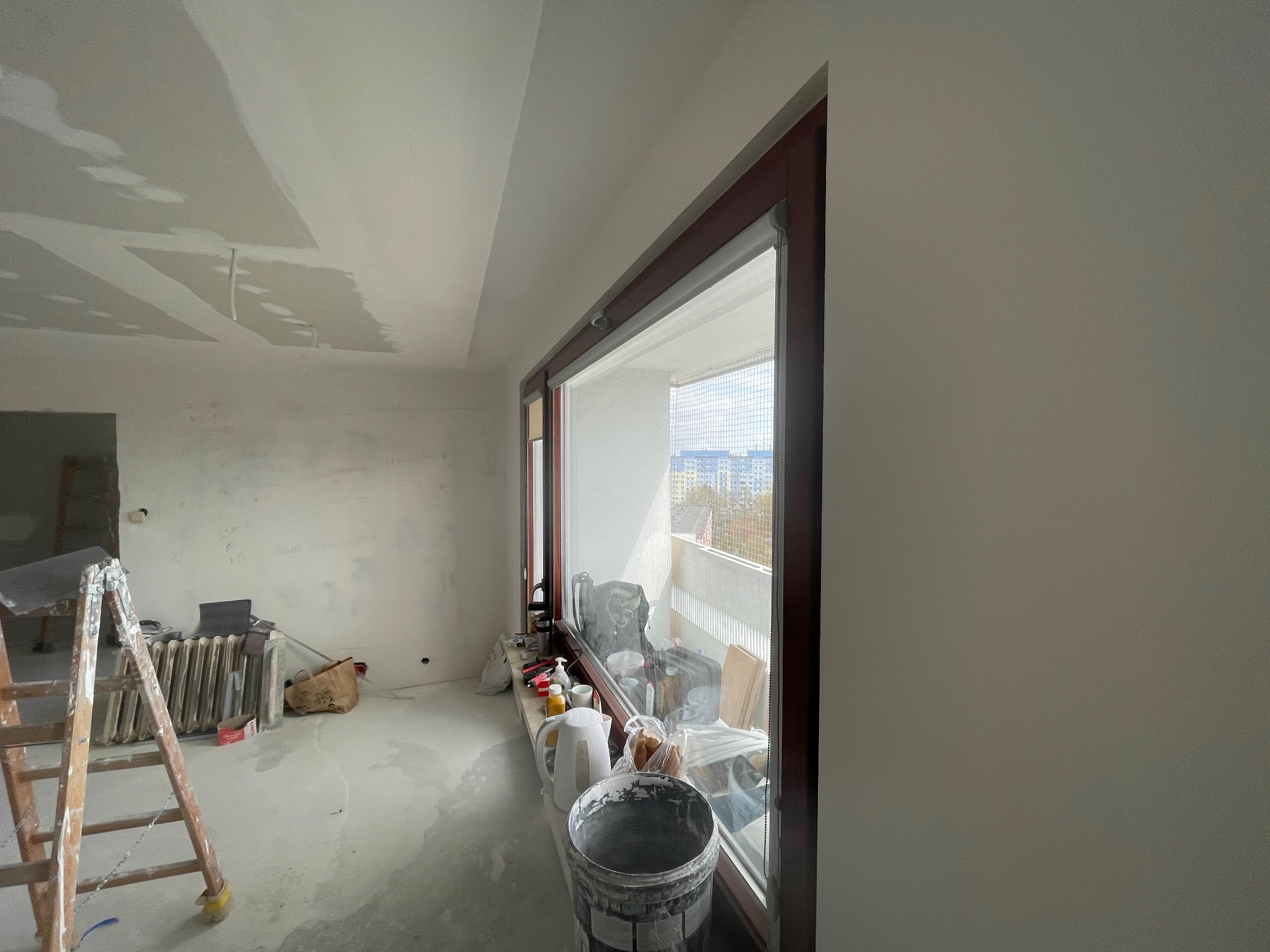 Pokój w trakcie remontu: ściany i sufit po szpachlowaniu, widoczne ślady po łataniu, drabina malarska, grzejnik i wiadro z resztkami farby przy oknie balkonowym.