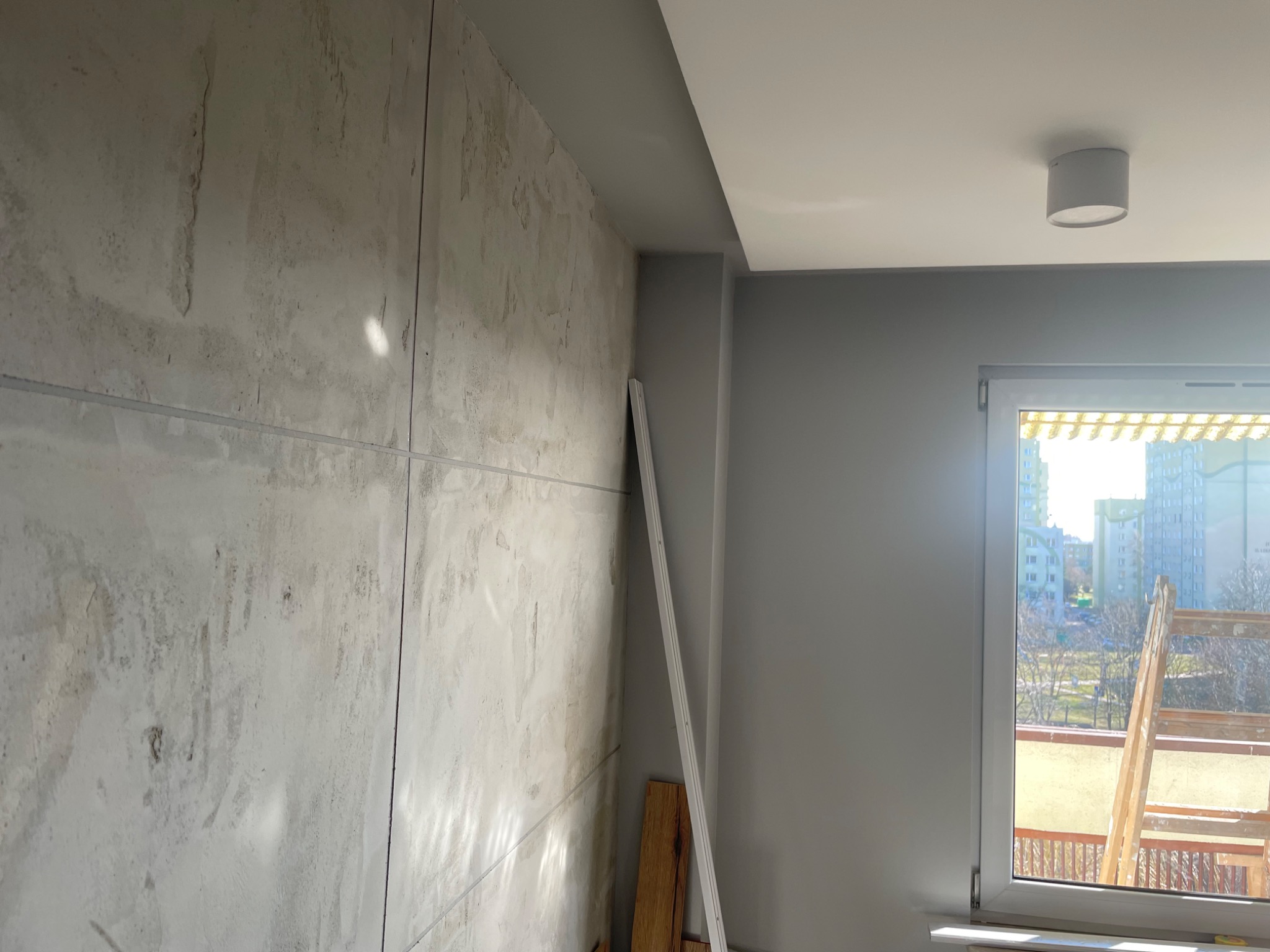 Surowa ściana z widocznymi śladami szpachlowania, panele gipsowe, przyścienna listwa i drabina oparta o okno z widokiem na osiedle mieszkaniowe.