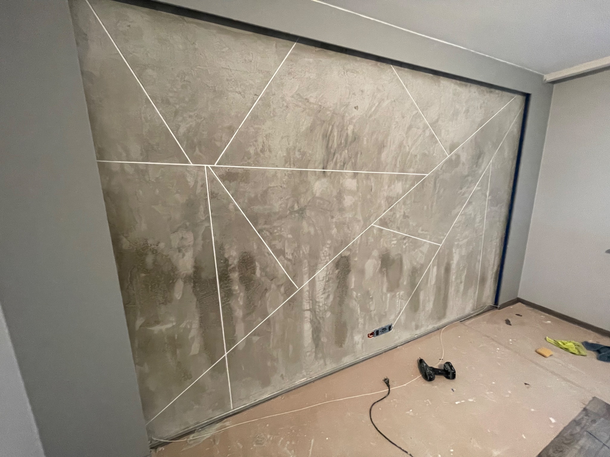 Ściana w pokoju po nałożeniu szarej, dekoracyjnej faktury z geometrycznym wzorem z białych linii. Widoczne gniazdko elektryczne, narzędzia i resztki materiałów na podłodze.