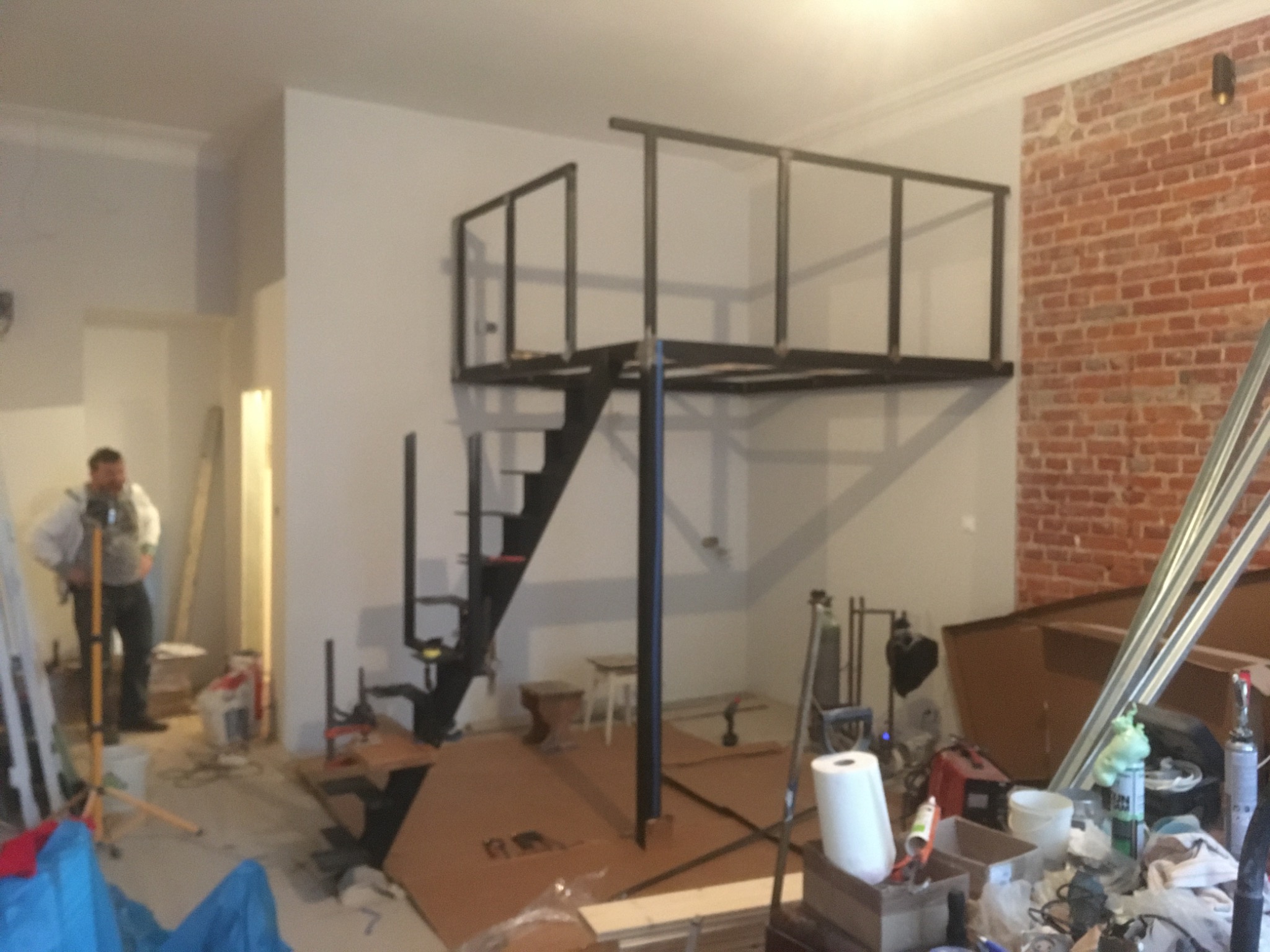 Wnętrze w trakcie remontu z metalową konstrukcją antresoli z czarnymi schodami, częściowo wykończona ściana z czerwonej cegły, widoczny pracownik w tle i narzędzia budowlane.