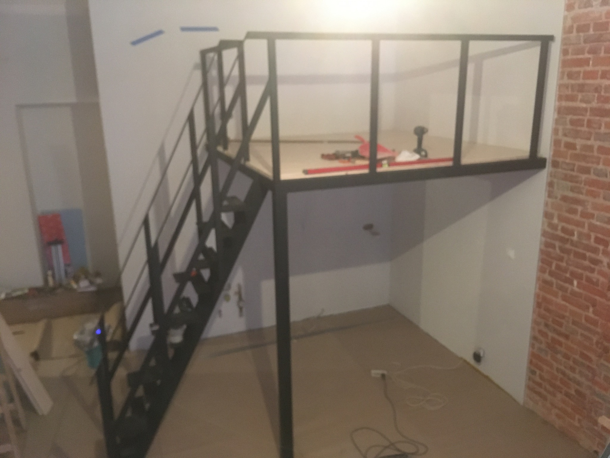 Metalowa konstrukcja antresoli z czarnymi schodami w surowym wnętrzu, widoczne narzędzia i materiały budowlane na platformie, ściana z czerwonej cegły w tle.