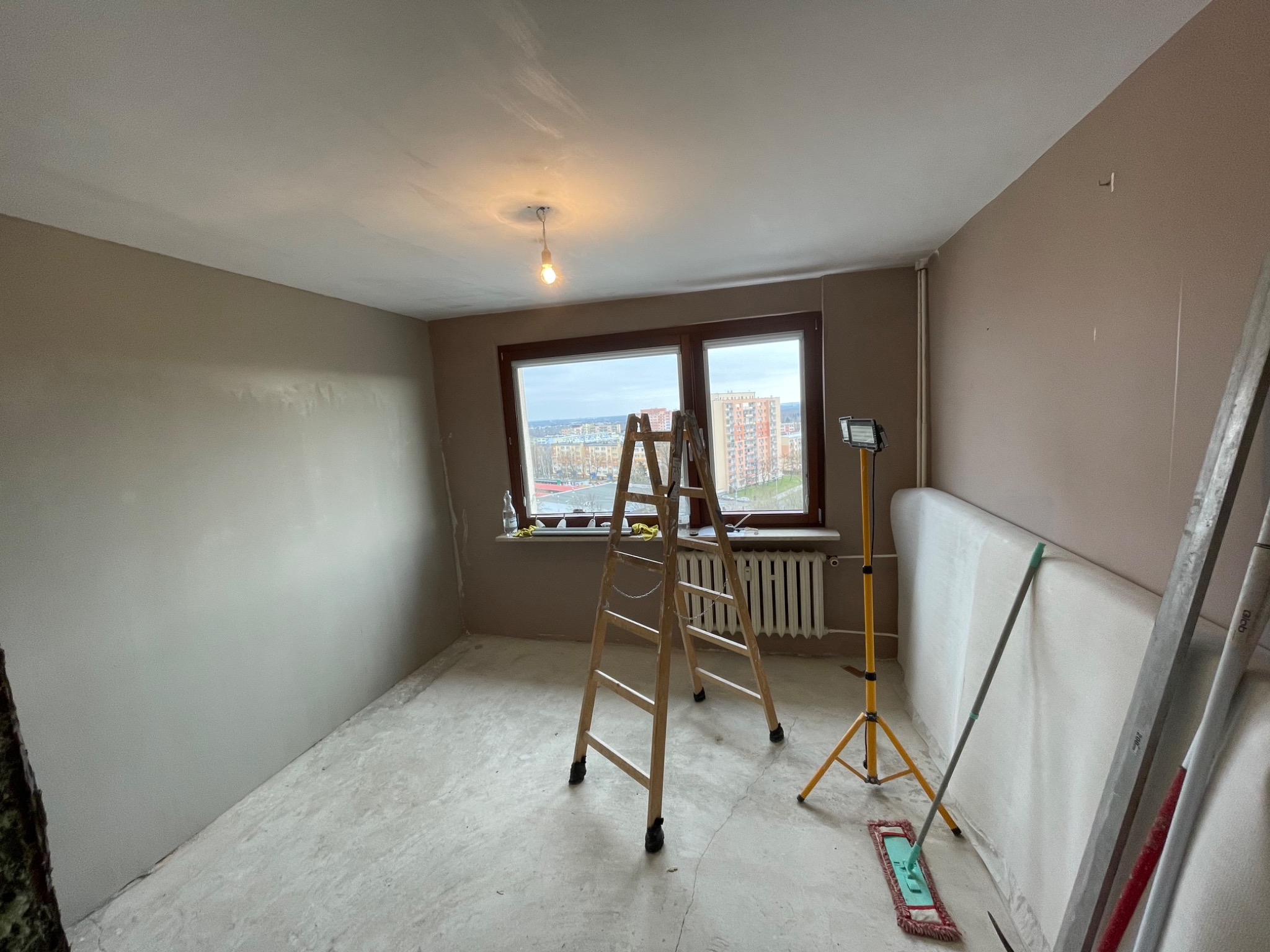 Pokój w trakcie remontu: świeżo wyszpachlowane ściany w odcieniach beżu i szarości, drewniana drabina stojąca przed oknem z widokiem na miasto, lampa budowlana na trójnogu, przygotowane płyty...