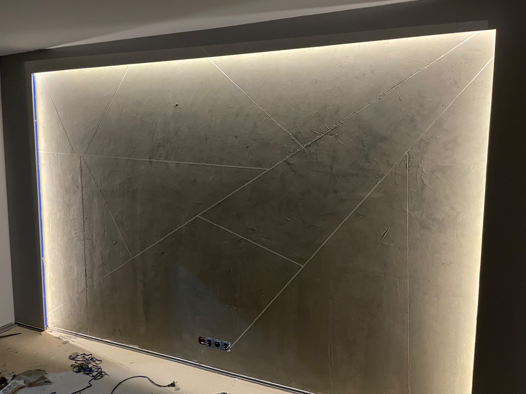 Ściana pokryta szpachlą dekoracyjną w geometryczne wzory, podświetlona listwami LED, widoczne gniazdka elektryczne i przewody na podłodze.