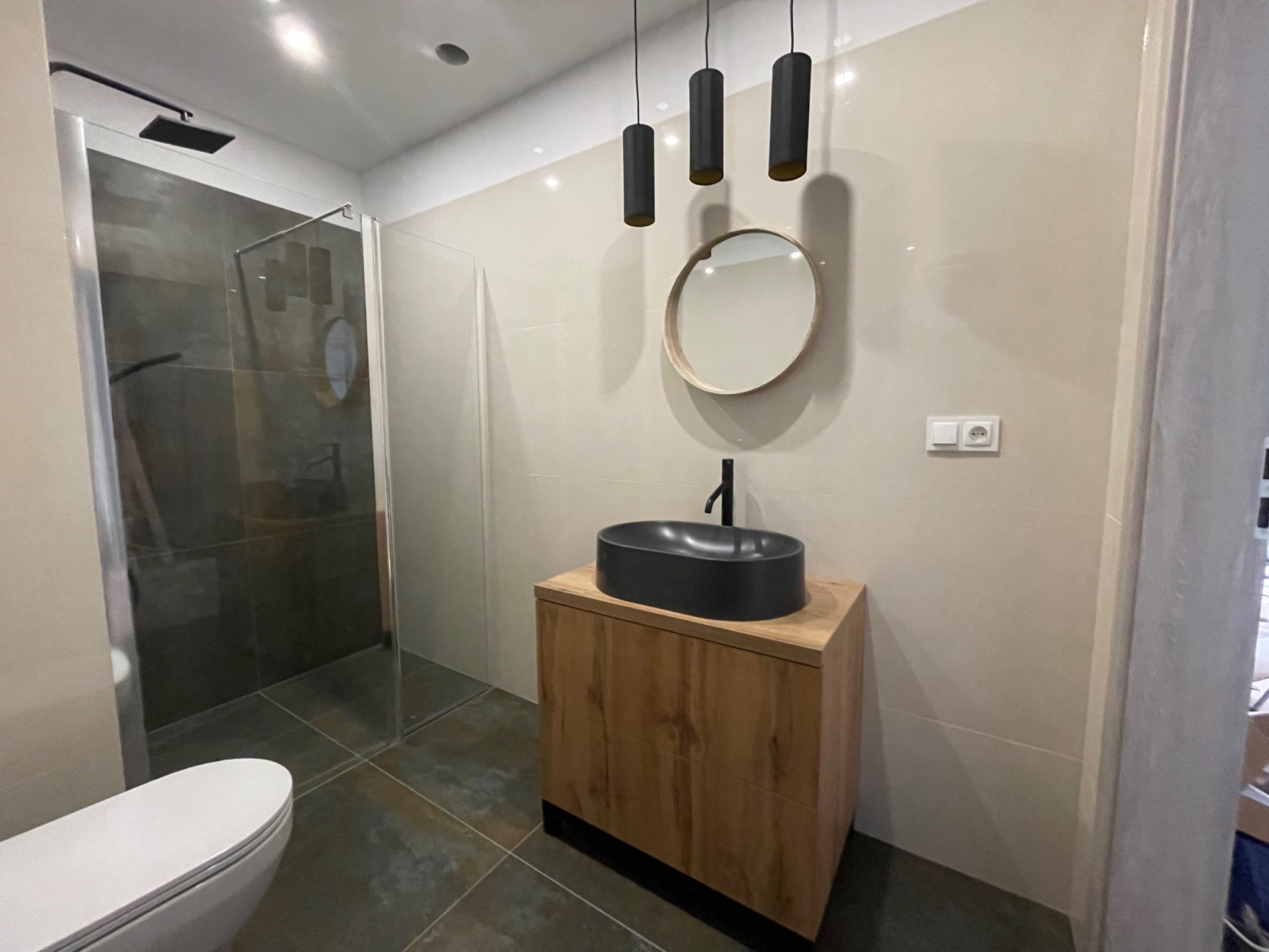Wykończona łazienka z kabiną prysznicową typu walk-in, umywalką nablatową na szafce z drewnopodobnym fornirem i okrągłym lustrem pod trzema cylindrycznymi lampami wiszącymi.