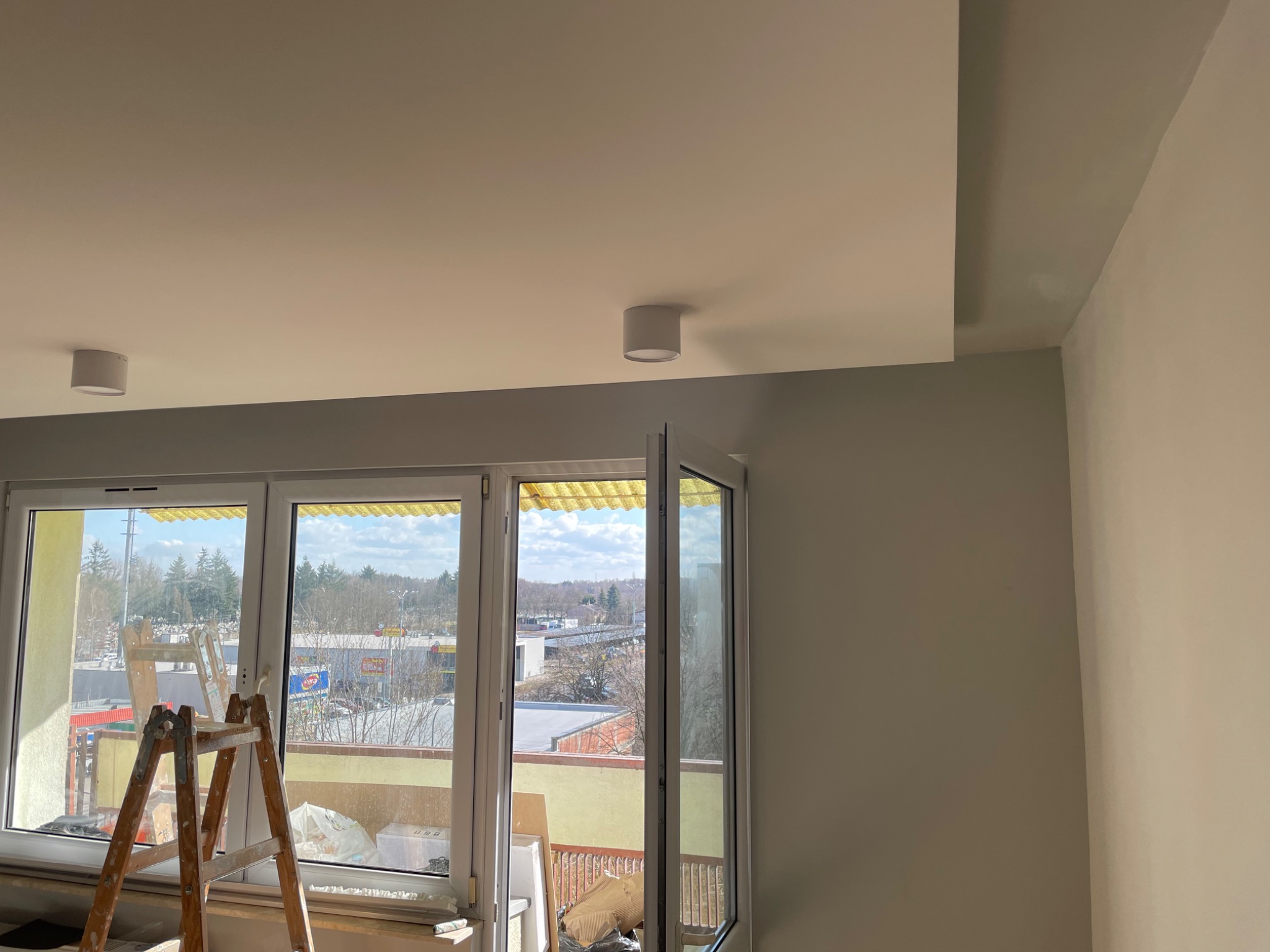 Wnętrze mieszkania w trakcie malowania, widoczna drabina przy oknie balkonowym, sufit pomalowany na biało, ściany w odcieniach szarości i beżu, zamontowane lampy sufitowe.