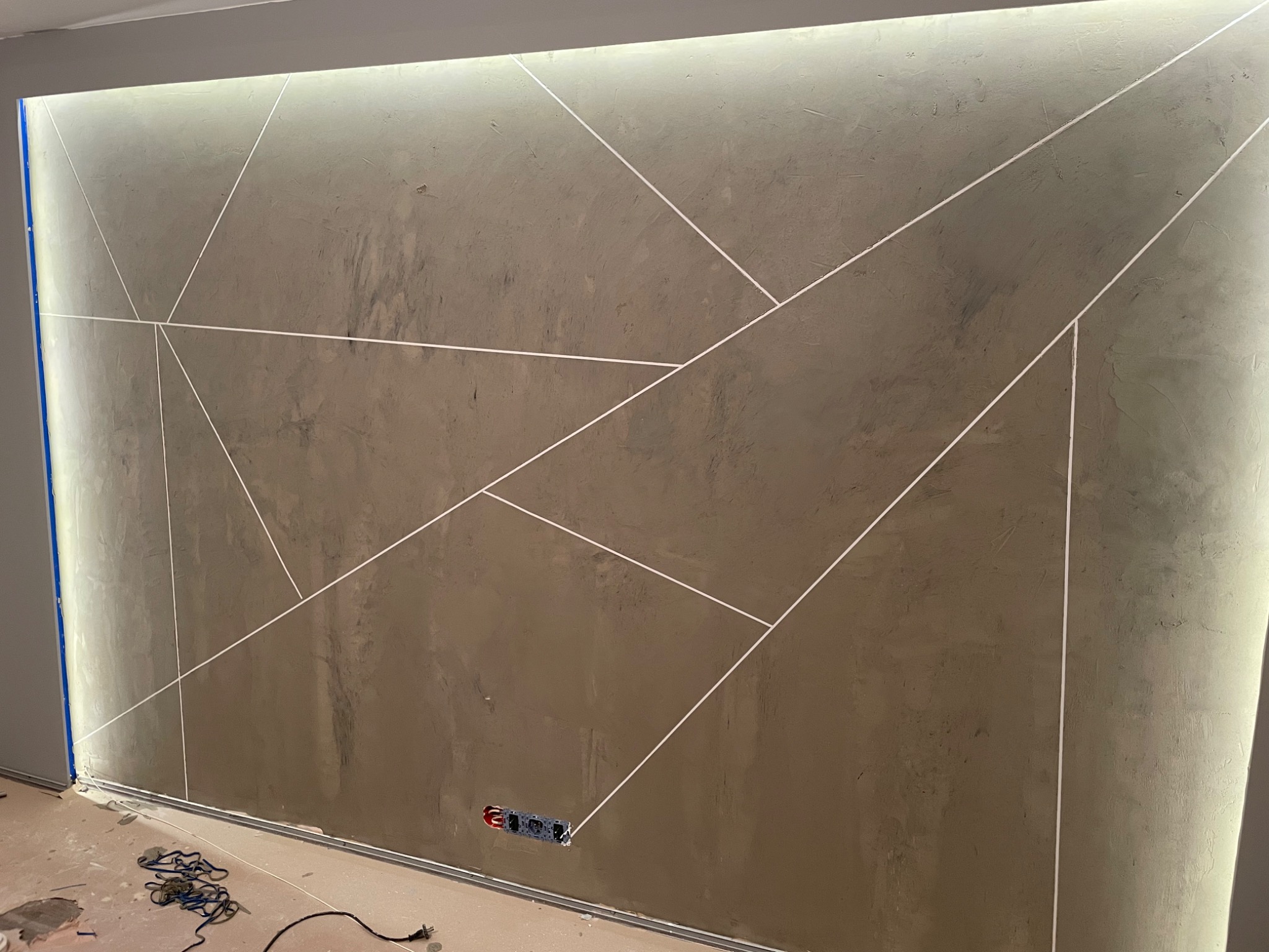 Ściana po szpachlowaniu z dekoracyjnymi liniami i podświetleniem LED w górnej części, widoczne gniazdko elektryczne i fragment podłogi w trakcie remontu.