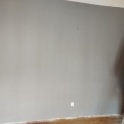 ADON Damian Klimek - Świeżo pomalowana szara ściana z widocznym gniazdkiem elektrycznym, fragmentem podłogi, drabiną i wiszącą kurtką roboczą w kolorze żółtym.