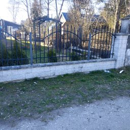 ADON Damian Klimek - Fragment ogrodzenia z murowaną podmurówką z szarej kostki i metalowym płotem z ozdobnymi elementami, widok na posesję z drzewami i zabudowaniami w tle.