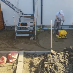 Pracownik ubija piasek zagęszczarką w przygotowaniu podłoża pod wylewkę, widoczne worki z cementem, łopaty, schody zewnętrzne i ogrodzenie.