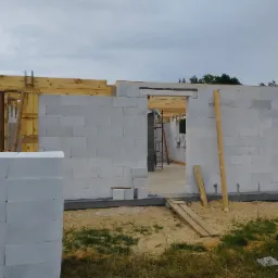 Wznoszenie ścian budynku z białych bloczków, widoczne drewniane szalunki i zbrojenie, paleta bloczków z napisem '24' w lewym dolnym rogu, teren budowy z piaskiem i elementami roślinności.