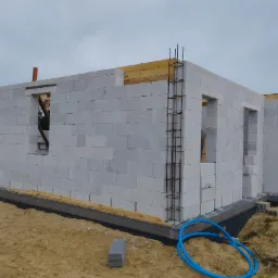 Wznoszenie ścian budynku z białych bloczk&oacute;w, widoczne otwory okienne i drzwiowe, zbrojenie narożnik&oacute;w, fundament zabezpieczony folią, niebieski wąż na piasku.