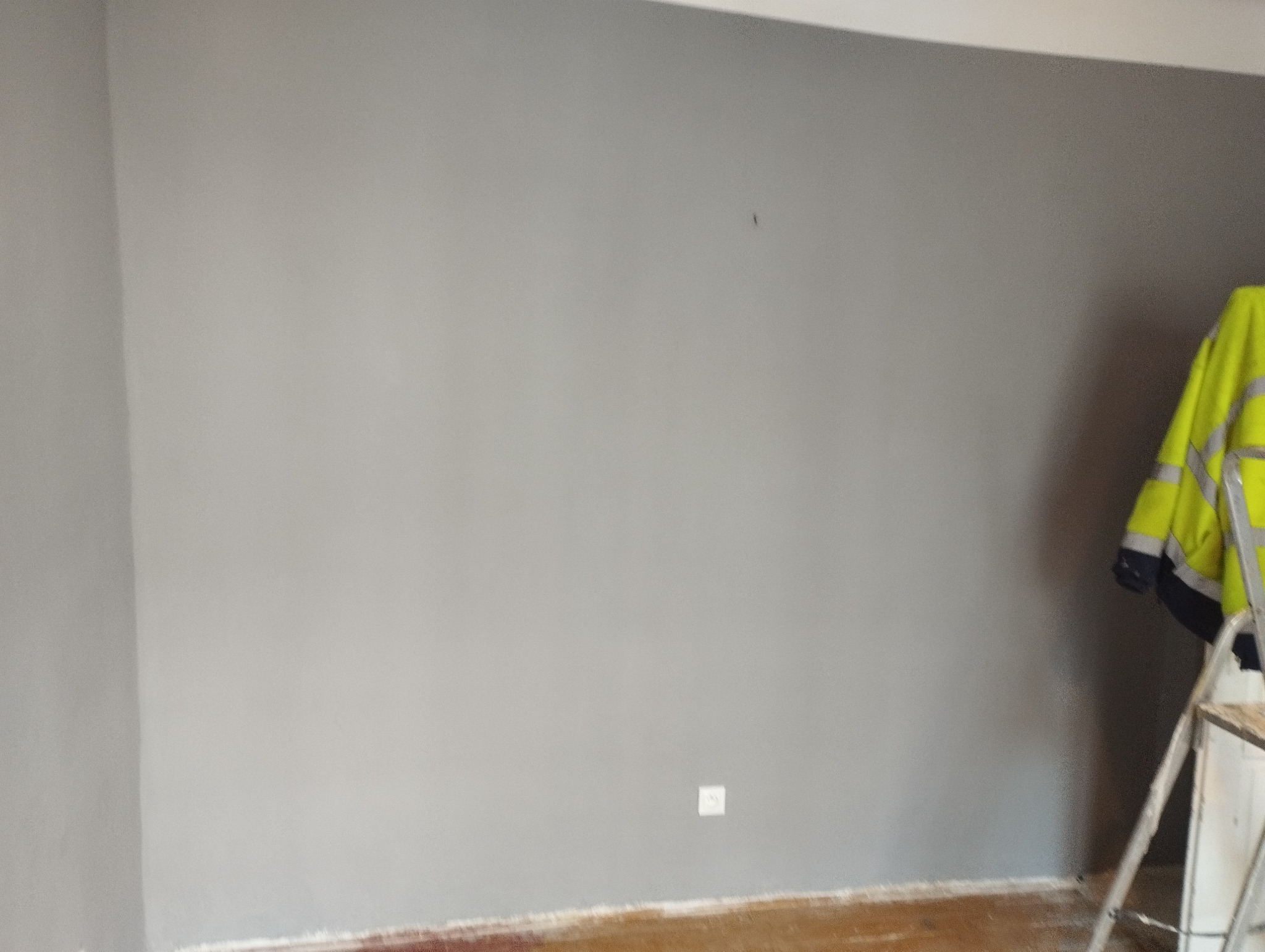 Świeżo pomalowana szara ściana z widocznym gniazdkiem elektrycznym, fragmentem podłogi, drabiną i wiszącą kurtką roboczą w kolorze żółtym.