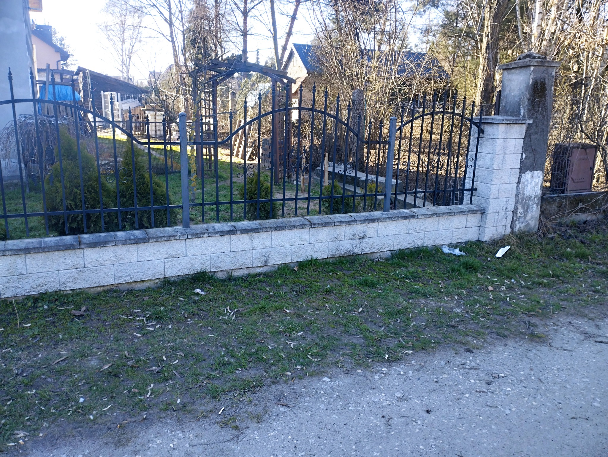 Fragment ogrodzenia z murowaną podmurówką z szarej kostki i metalowym płotem z ozdobnymi elementami, widok na posesję z drzewami i zabudowaniami w tle.