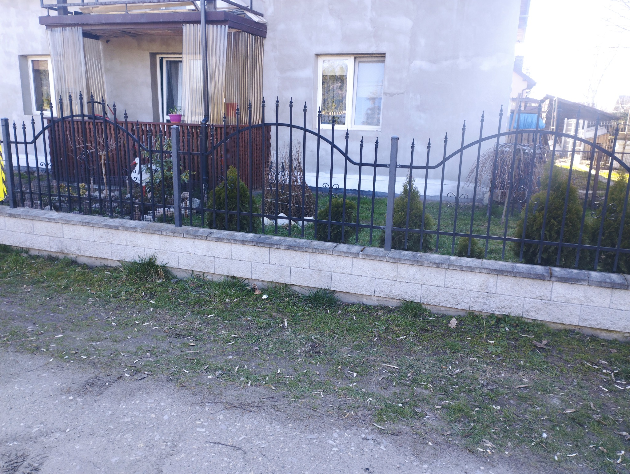 Szary dom jednorodzinny z czarnym, metalowym ogrodzeniem na niskiej, murowanej podmurówce, z widocznymi młodymi tujami posadzonymi wzdłuż ogrodzenia.