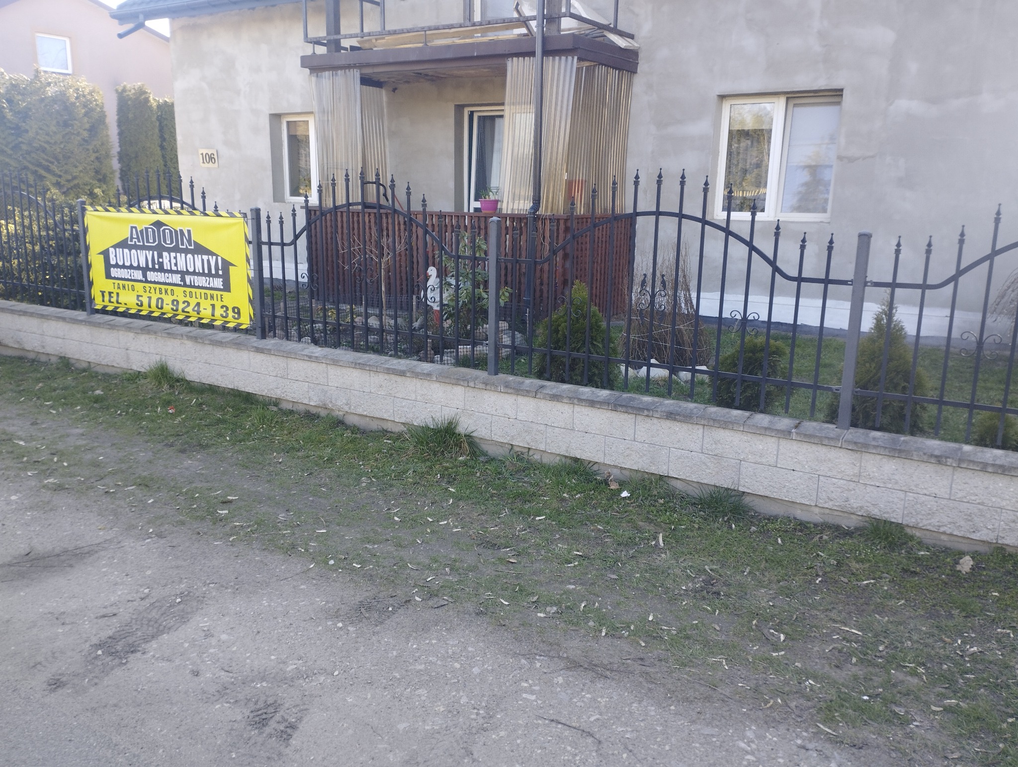 Żółto-czarny baner reklamowy firmy budowlanej i remontowej 'ADON' z numerem telefonu, przymocowany do metalowego ogrodzenia przed domem z szarym tynkiem i balkonem.