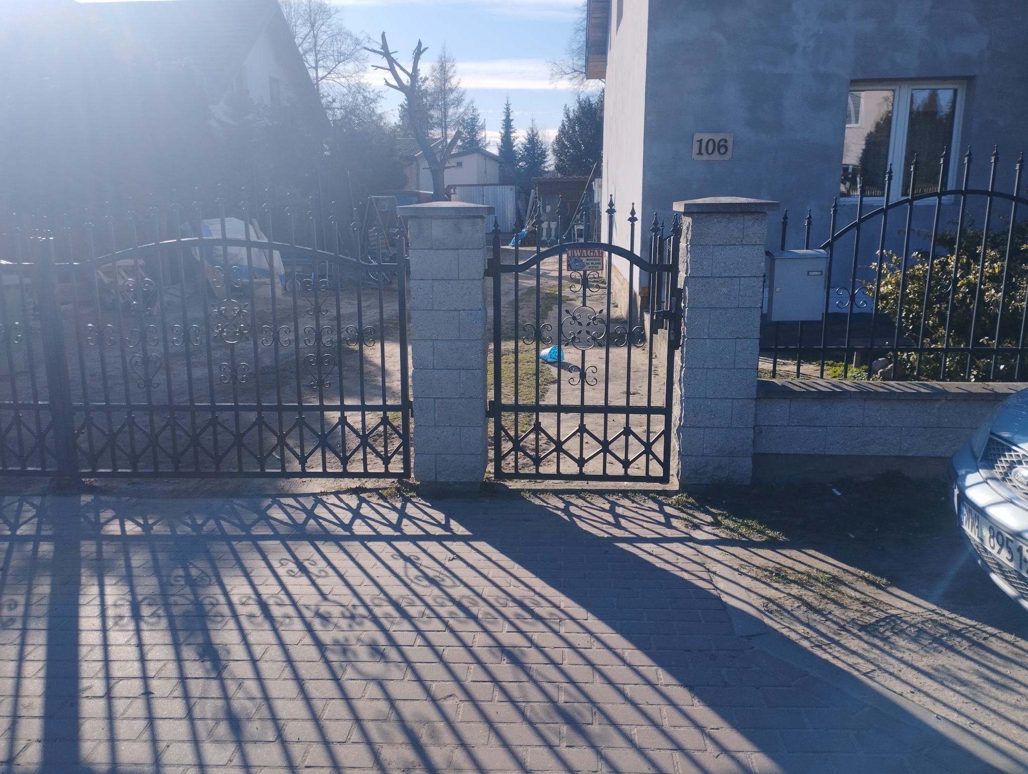 Otwarta, czarna, metalowa brama wjazdowa z dekoracyjnymi elementami, osadzona na słupkach z szarej kostki, rzucająca długie cienie na brukowany podjazd przed domem z numerem 106.
