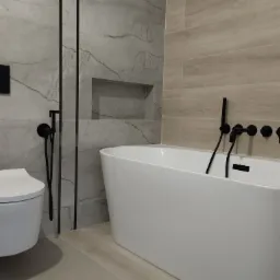 Nowoczesna łazienka z płytkami imitującymi marmur, wiszącą toaletą z czarnym przyciskiem, białą wanną wolnostojącą i czarną armaturą.
