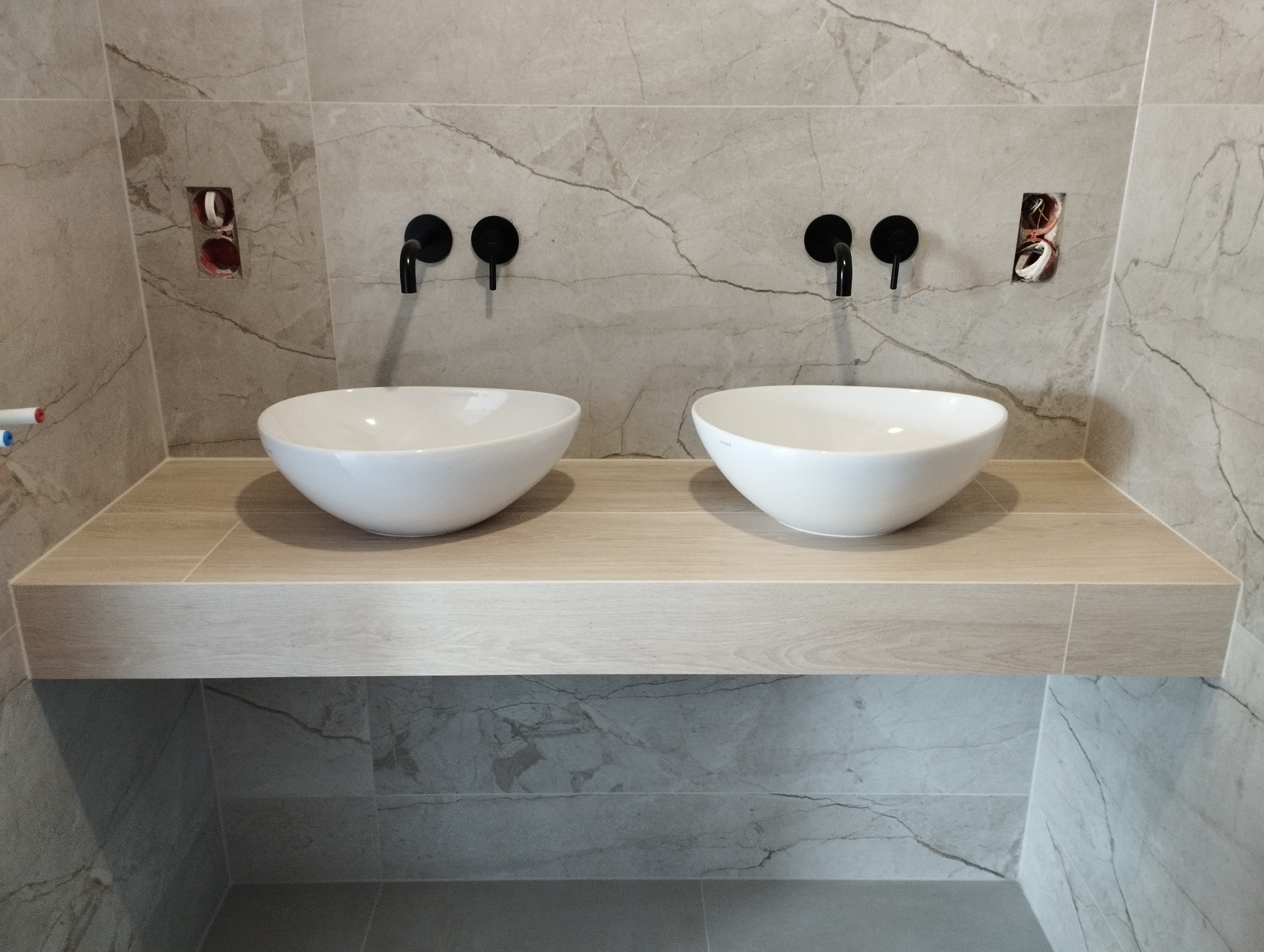 Dwie owalne, białe umywalki nablatowe z czarnymi bateriami, zamontowane na drewnopodobnym blacie z płytek, na tle marmurowych płytek ściennych; widoczne niedokończone otwory na gniazdka.