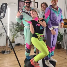 Grupa czterech os&oacute;b w strojach inspirowanych latami 80. pozuje na obrotowej platformie fotobudki 360, otoczonej bańkami mydlanymi i oświetleniem LED.