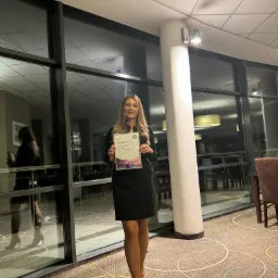 Kobieta w czarnej sukience trzyma certyfikat przed oknem w eleganckim wnętrzu z wzorzystą wykładziną.