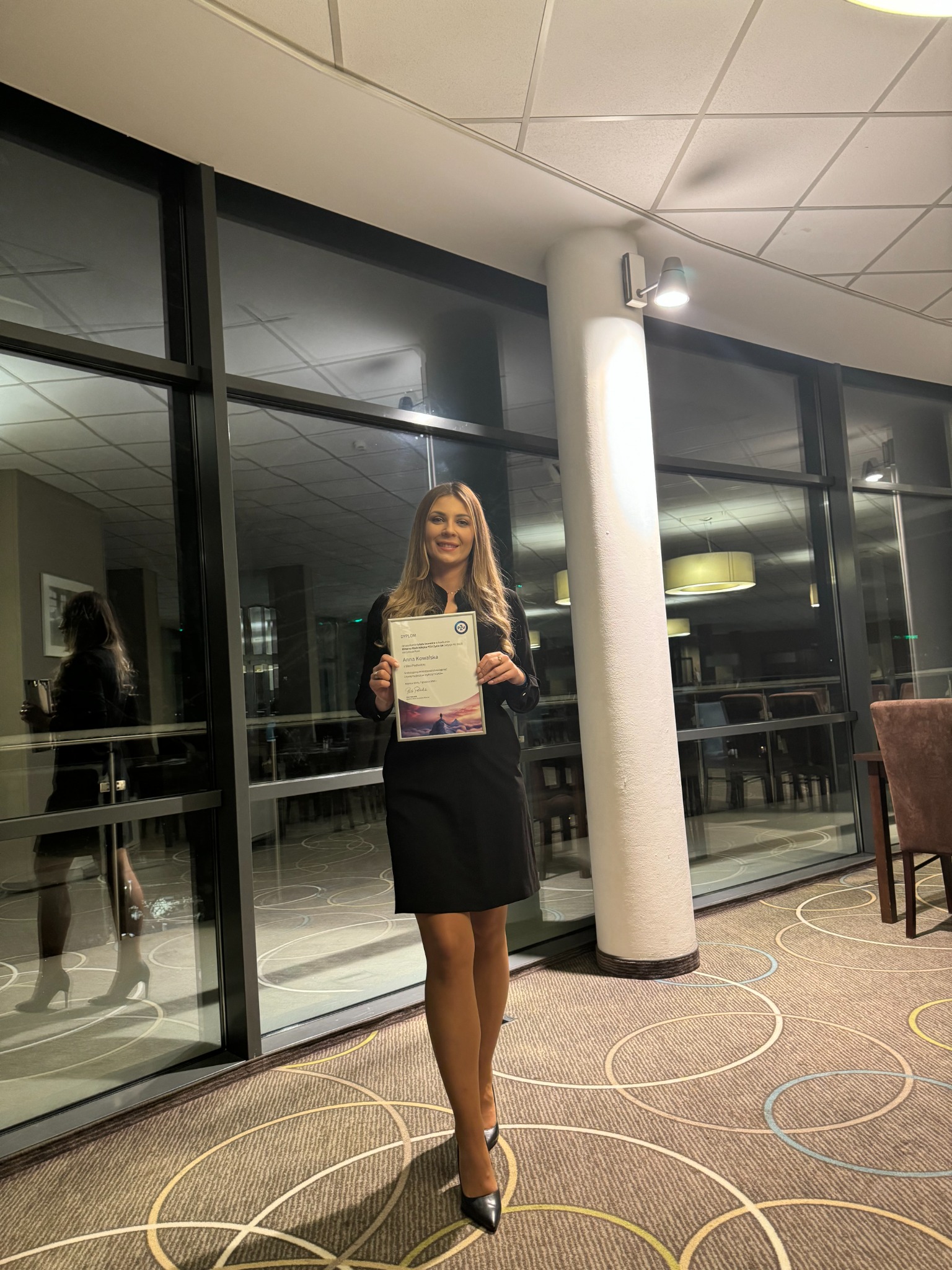 Kobieta w czarnej sukience trzyma certyfikat przed oknem w eleganckim wnętrzu z wzorzystą wykładziną.