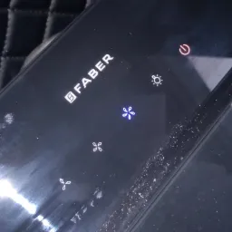 Czarny panel sterowania okapu kuchennego marki Faber z podświetlanymi ikonami funkcji, widoczne ślady użytkowania i drobne zabrudzenia.