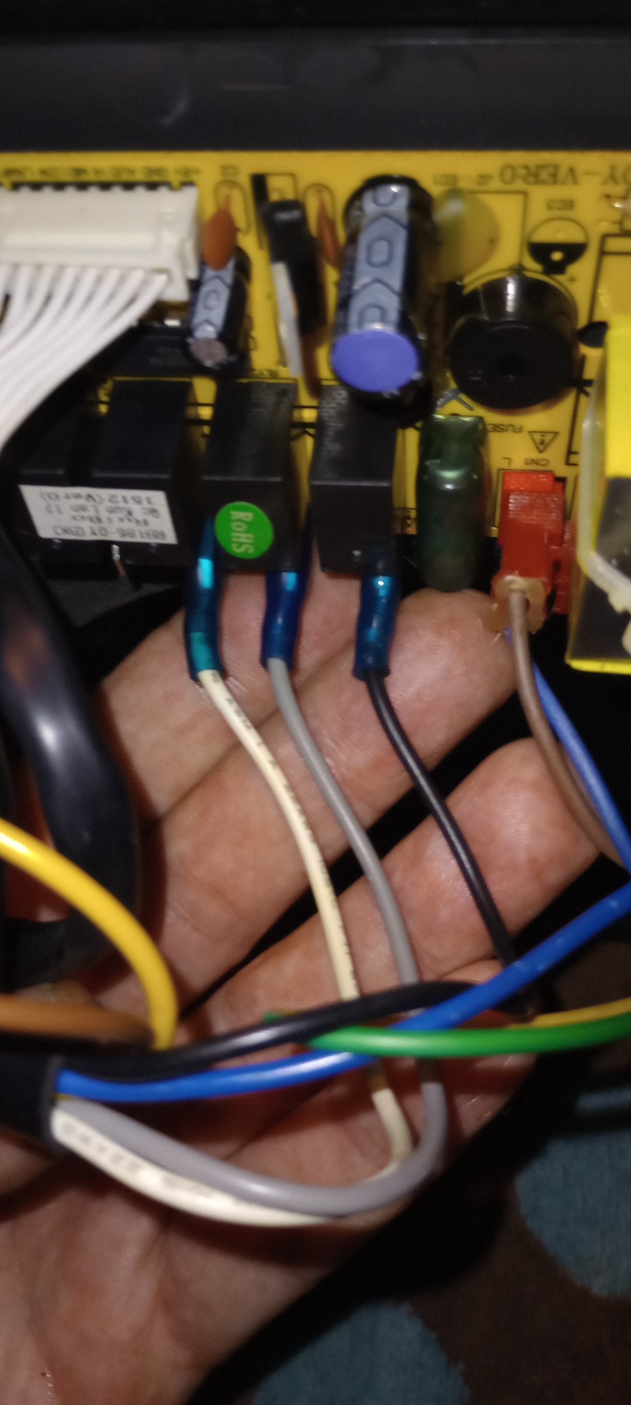 Zbliżenie na dłoń trzymającą przewody elektryczne podłączone do płytki drukowanej z elementami elektronicznymi, w tym czarnymi przekaźnikami z zieloną naklejką RoHS.