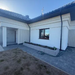 Elewacja domu jednorodzinnego w jasnych barwach z elementami dekoracyjnymi. Widoczne wykończone ściany, okna i kostka brukowa na podjeździe.