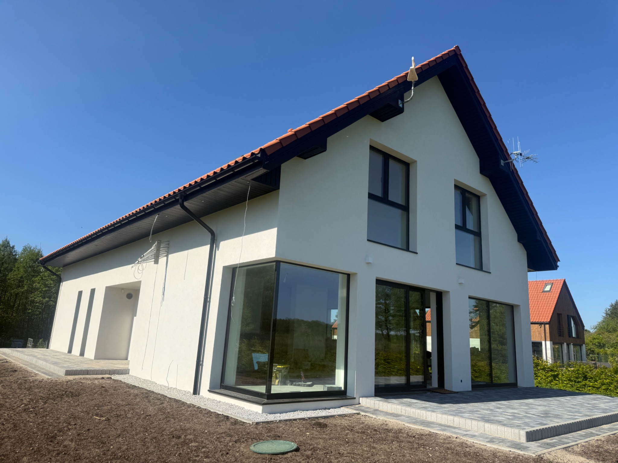Nowoczesny dom jednorodzinny z białymi ścianami i dużymi oknami w Ełku, widok z zewnątrz, błękitne niebo w tle. Czerwona dachówka i czarne obramowania okien.
