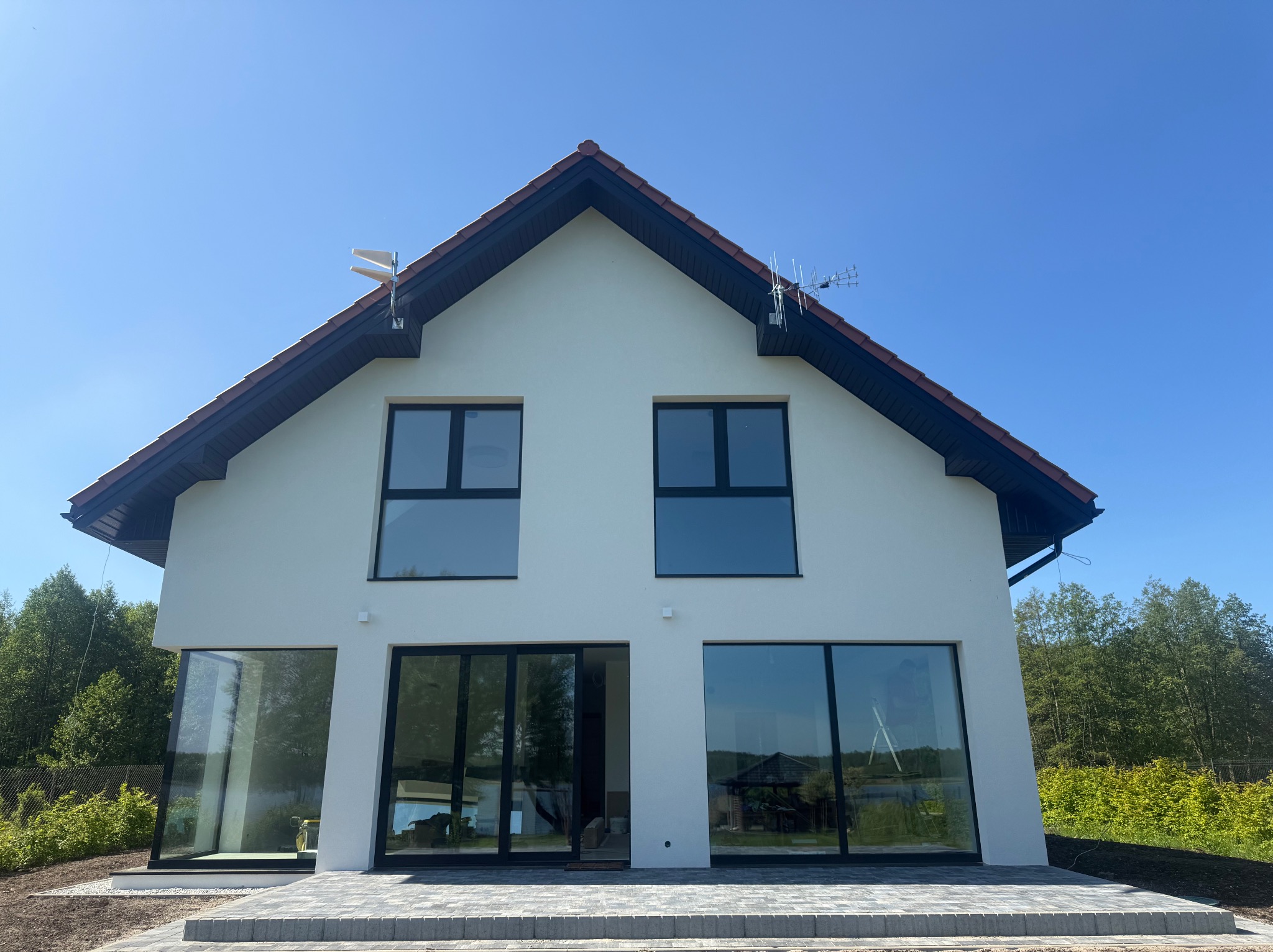 Nowoczesny, dwukondygnacyjny dom z białymi ścianami i dużymi oknami, otoczony zielenią. Widoczne detale wykończenia elewacji i zagospodarowania terenu.