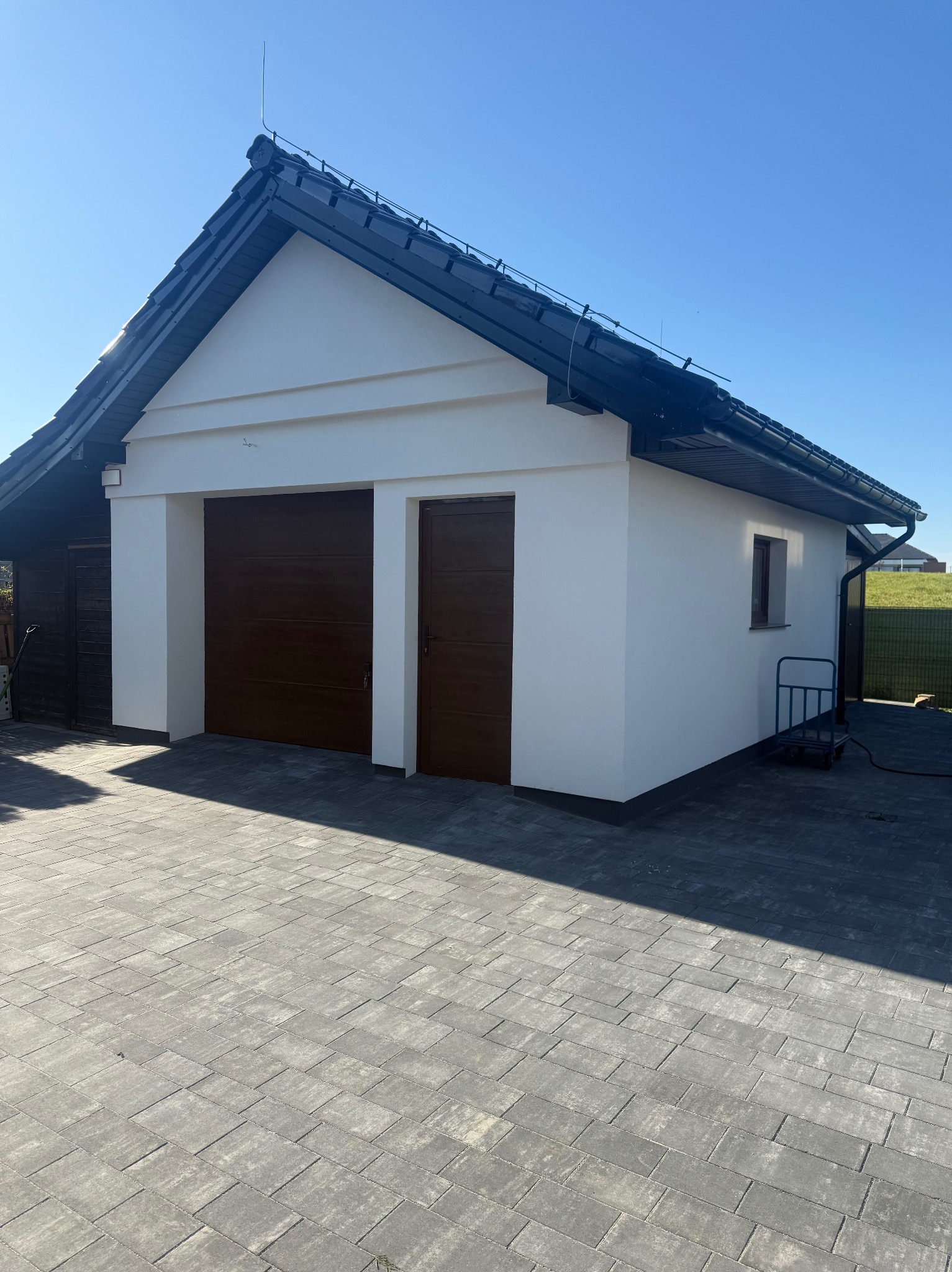 Biały budynek z brązowymi bramami garażowymi i drzwiami, czarny dach, kostka brukowa na podjeździe, błękitne niebo. Widok na elewację boczną i frontową.