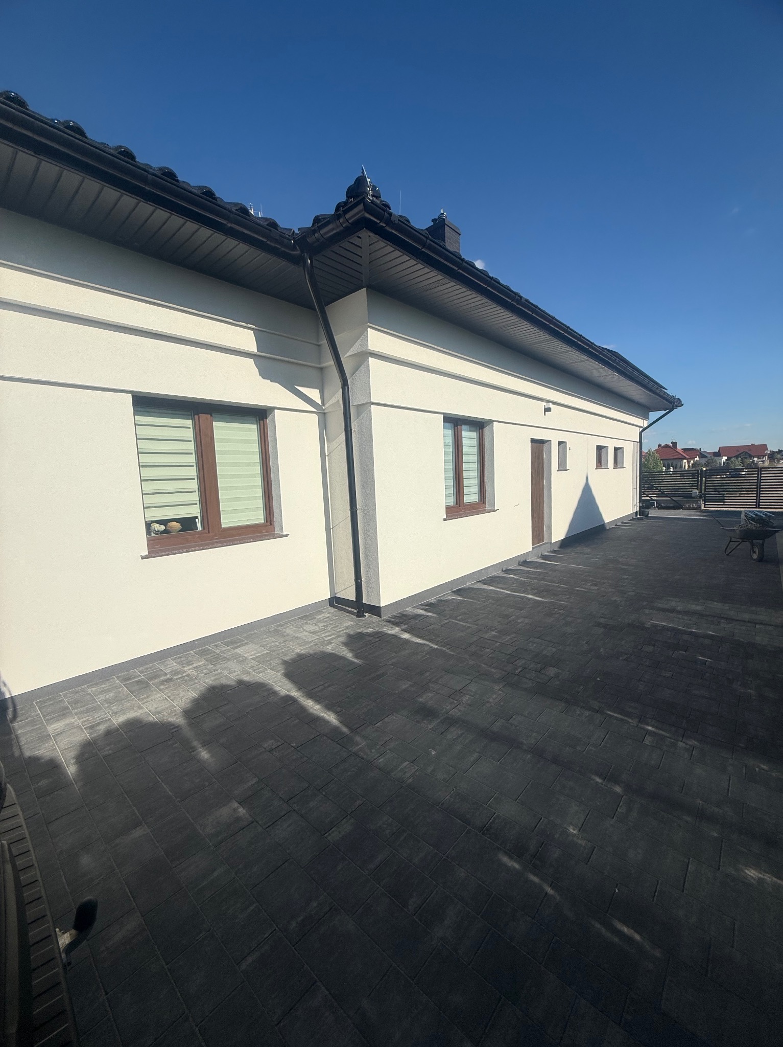 Biała elewacja domu jednorodzinnego z brązowymi ramami okiennymi i antracytową kostką brukową na podjeździe w słoneczny dzień. Widok z dołu.