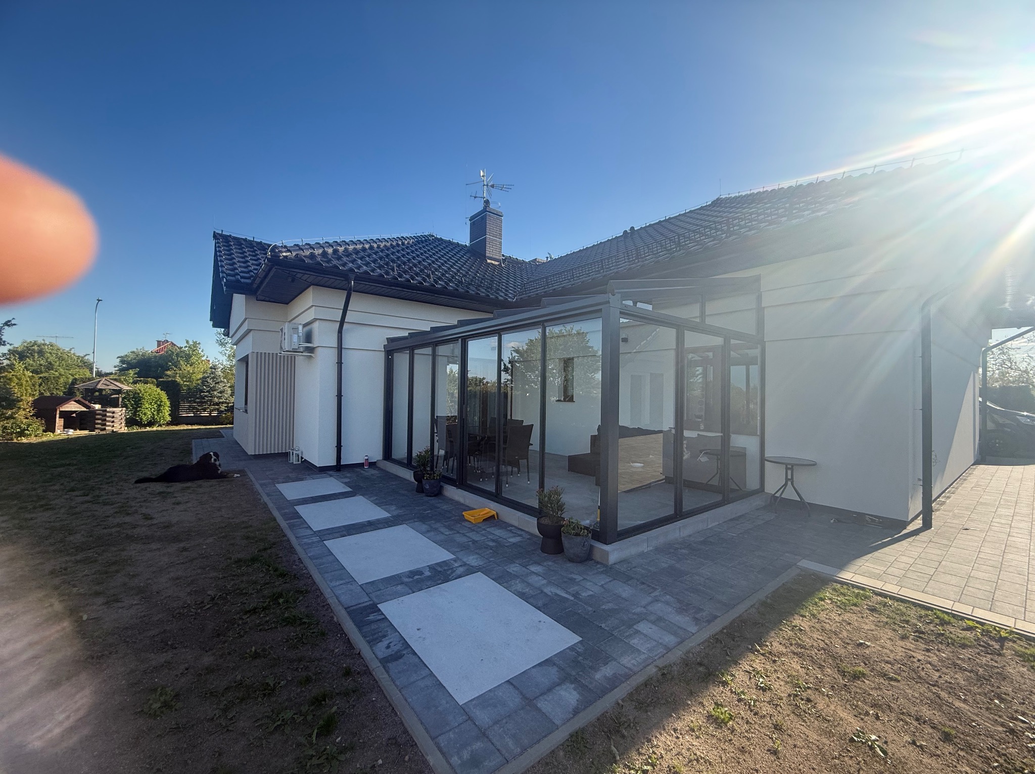 Biały dom z przeszkloną oranżerią i kostką brukową. Na trawniku leży pies. Słoneczny dzień, błękitne niebo.