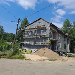 Dwukondygnacyjny dom w trakcie prac elewacyjnych, z widocznym rusztowaniem, częściowo ocieplony szarymi płytami izolacyjnymi, z odsłoniętą cegłą na bocznej ścianie i stertą piasku przed budynkiem