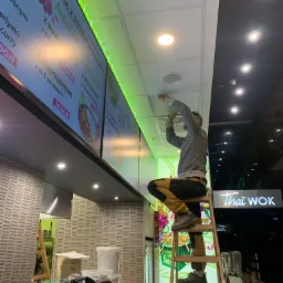 Mężczyzna na drabinie montuje lub naprawia głośnik sufitowy w lokalu gastronomicznym Thai Wok, widoczne menu elektroniczne z cenami dań.