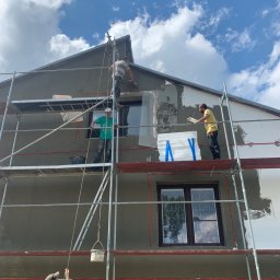 AllMar Marcin Moczulski - Elewacja domu w trakcie szpachlowania i ocieplania, widoczne rusztowania, pracownicy w trakcie pracy, styropianowe płyty z niebieską taśmą, szpachlowana ściana pod błękitnym niebem.