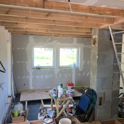 AllMar Marcin Moczulski - Wnętrze w trakcie prac wykończeniowych: ściany po szpachlowaniu z widocznymi łatami, drewniana konstrukcja sufitu, narzędzia i materiały budowlane na podłodze, drabina oparta o komin z betonu...