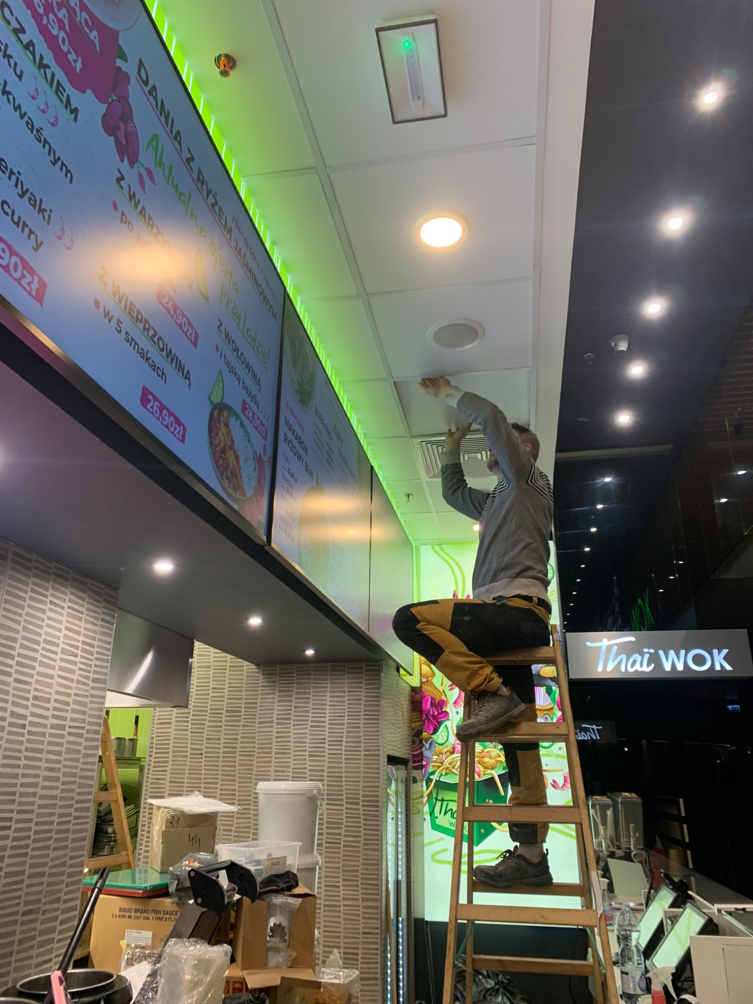 Mężczyzna na drabinie montuje lub naprawia głośnik sufitowy w lokalu gastronomicznym Thai Wok, widoczne menu elektroniczne z cenami dań.