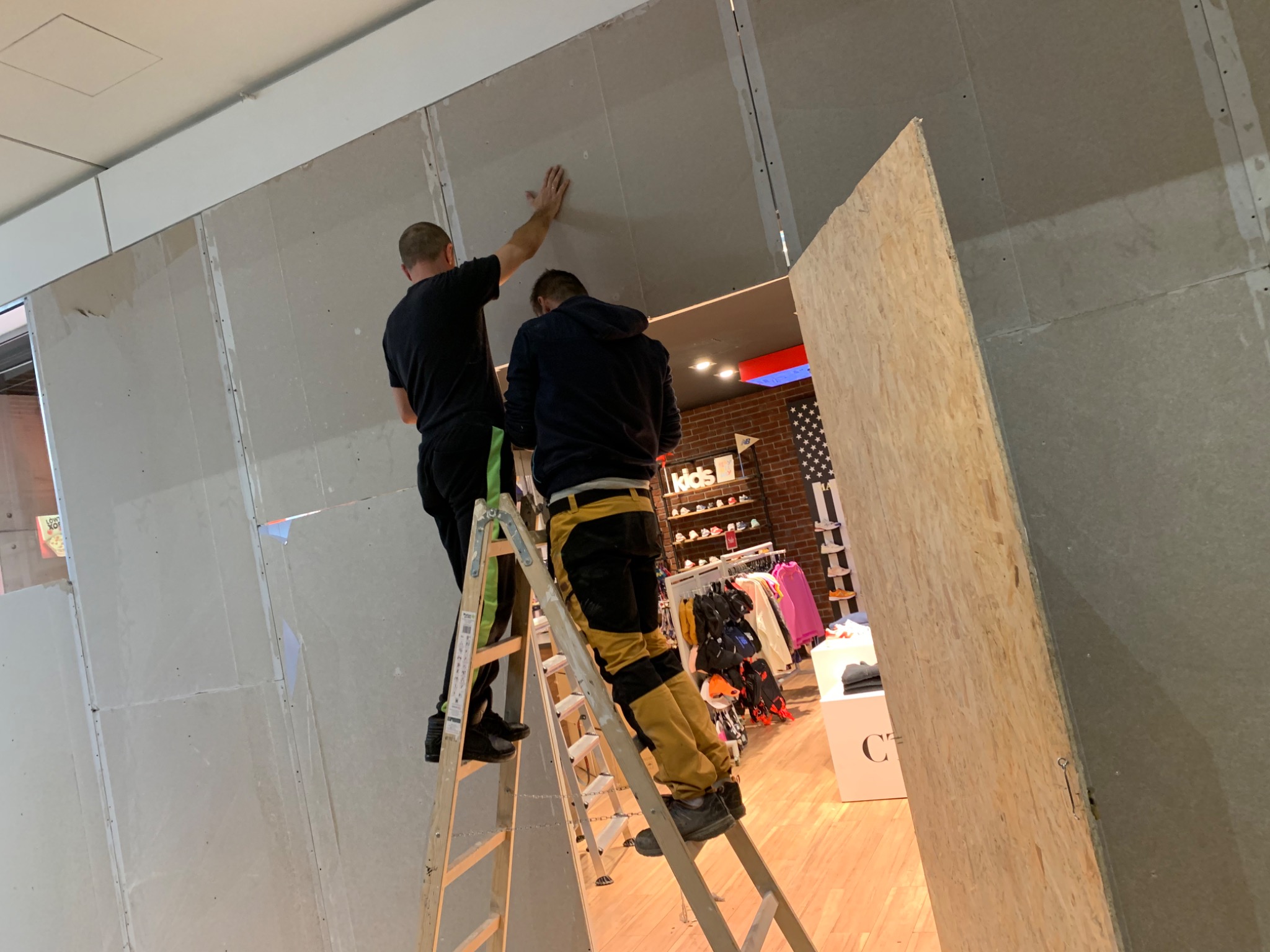 Dwóch mężczyzn na drabinach montuje płyty kartonowo-gipsowe wewnątrz lokalu handlowego z widocznym asortymentem odzieżowym w tle.