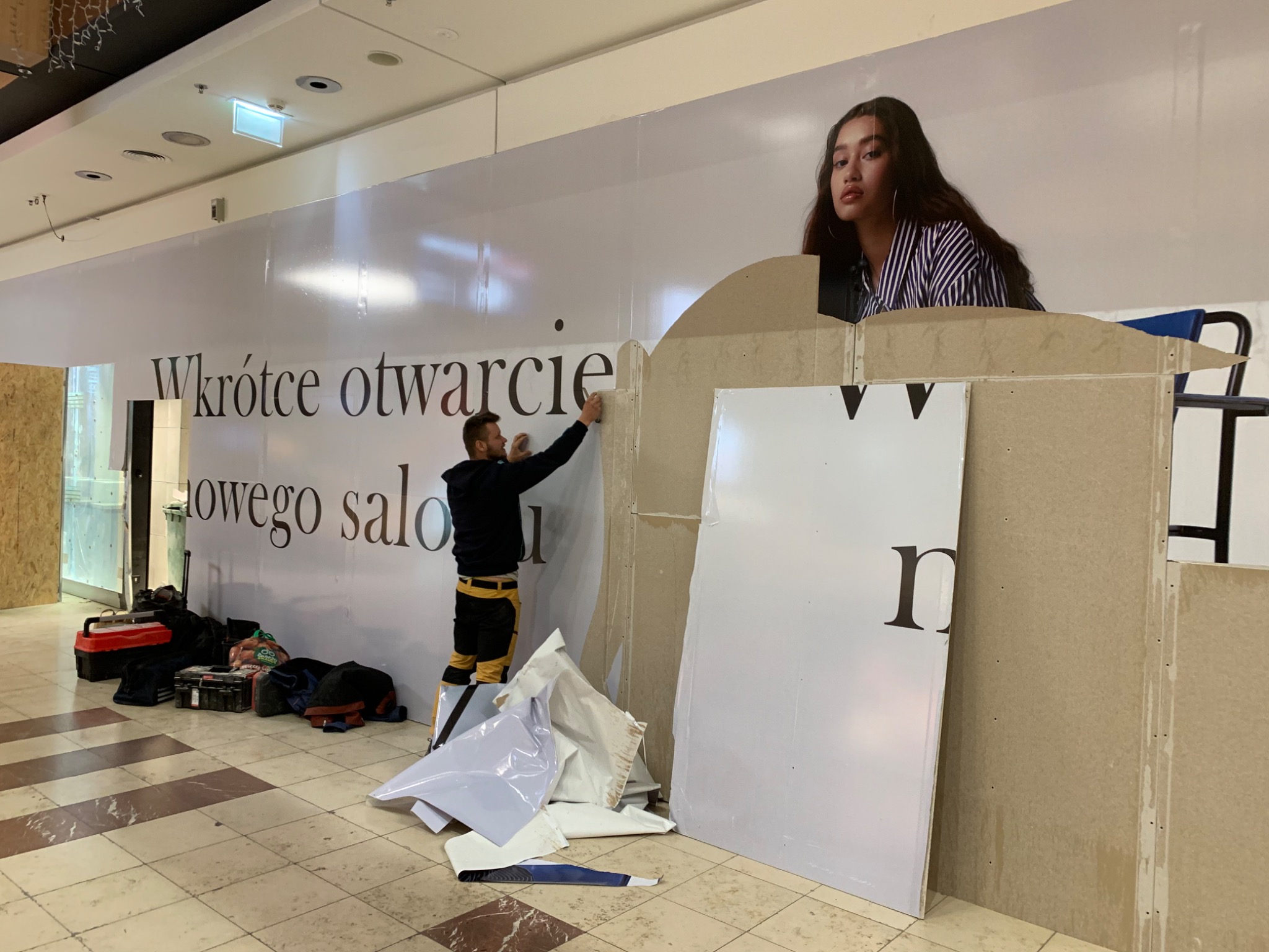 Pracownik na drabinie montuje duży baner reklamowy z napisem 'Wkrótce otwarcie nowego salonu' i wizerunkiem kobiety w Legionowie. Na podłodze leżą narzędzia i resztki materiałów.