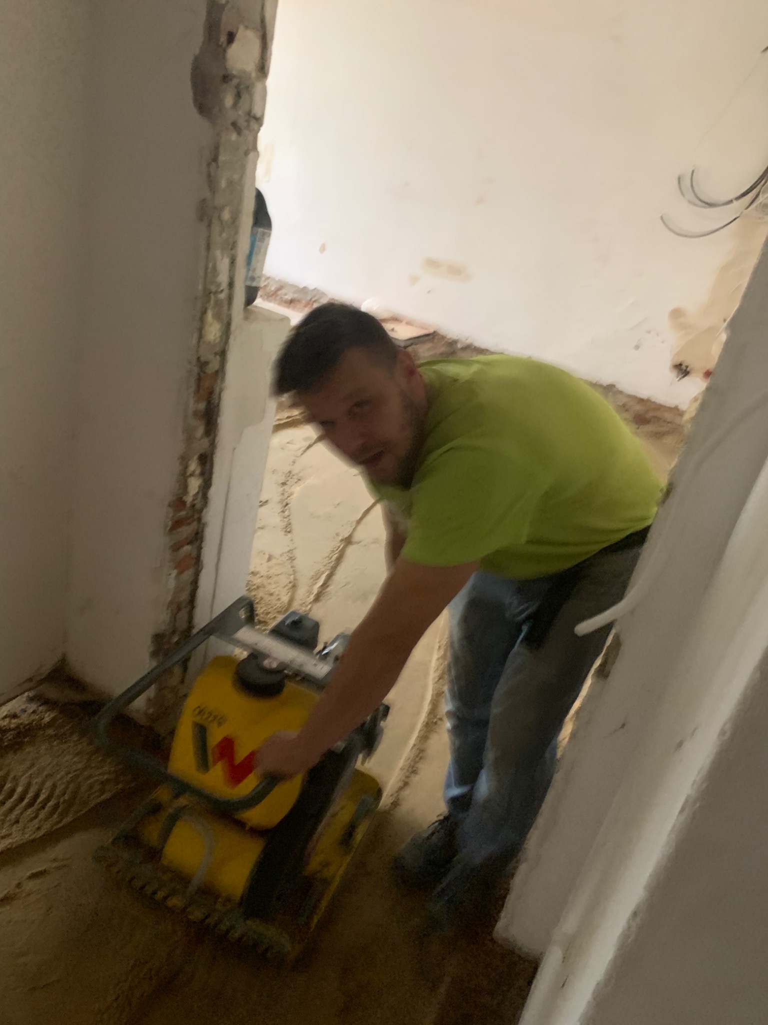 Mężczyzna w zielonej koszulce ubija podłoże żółtą zagęszczarką w trakcie prac przygotowawczych do remontu mieszkania, widoczne fragmenty ścian w stanie surowym.